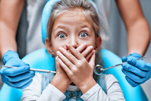 Close up view of a little girl looking scared and terrified screaming covering her mouth from the dentists with medical tools as a result of dental anxiety