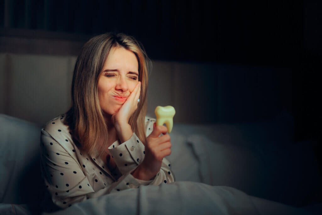 Woman in polka dot pajamas holding a plastic tooth, touching cheek with pained expression, suggesting toothache.